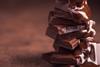 A close-up photograph of melted chocolate being poured over a pile of chocolate pieces against a light brown background