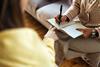 Two people sit together talking, as one person gestures towards a notebook and the other writes inside it