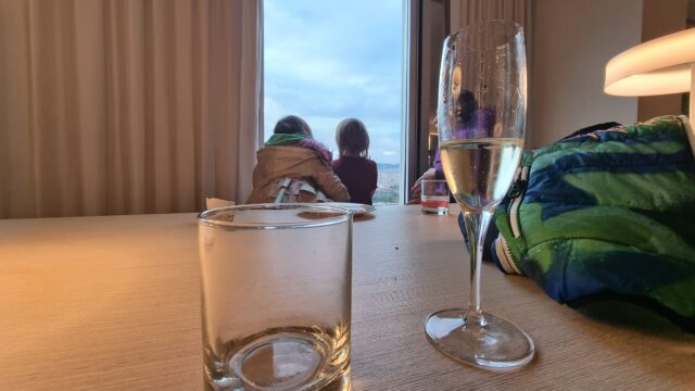 Half-full champagne flute on a table. In the background, two children look out of a floor-to-ceiling window.