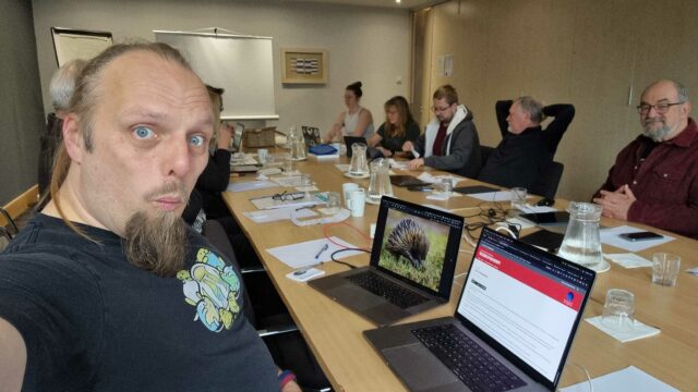 Dan sits in front of two laptops (one of which shows a photo of an echidna for some reason), in a meeting room full of casually-dressed volunteers.