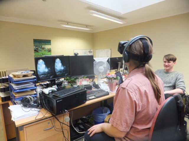 Dan, wearing a VR headset, sits in an office environment, watched by Adam.