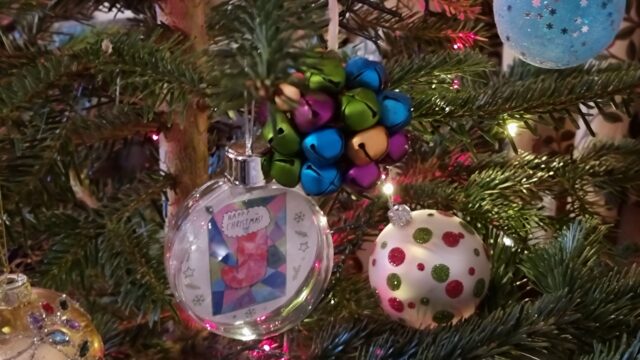 A cluster of three baubles hangs among pink and white fairy lights: one is a multicoloured assortment of bells, another is a plain white bauble decorated with glittery green and red spots, a third is a transparent plastic sphere containing a colourful children's drawing of a stocking.