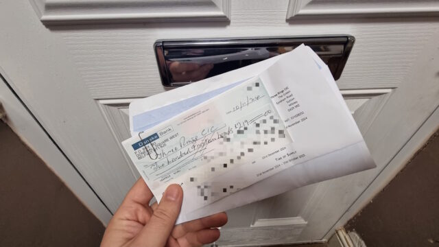 In front of a letterbox, an hand holds an open envelope and a (censored) Danske Bank cheque for £219.