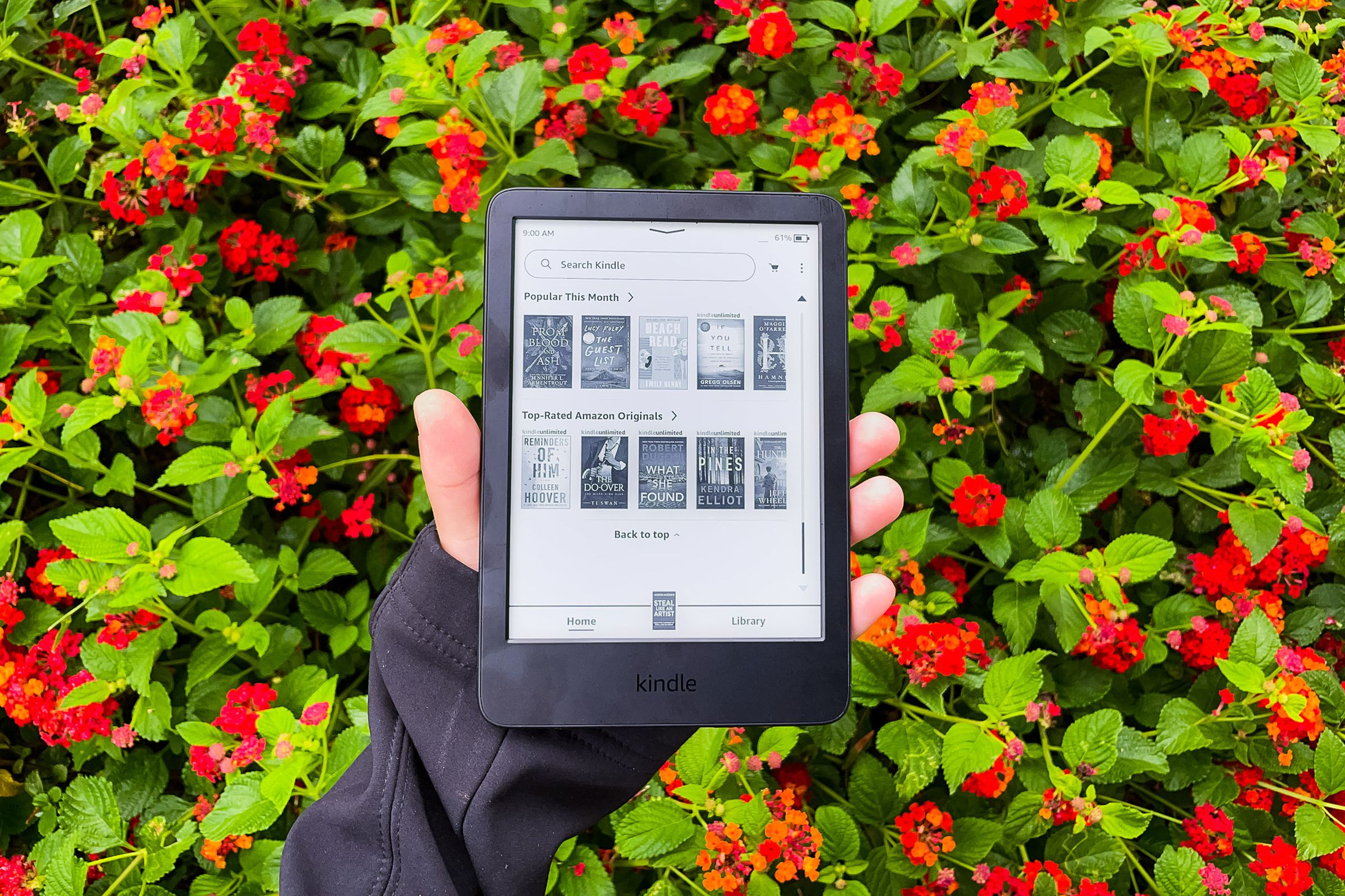 A hand holding the 2022 Kindle in front of red flowers.