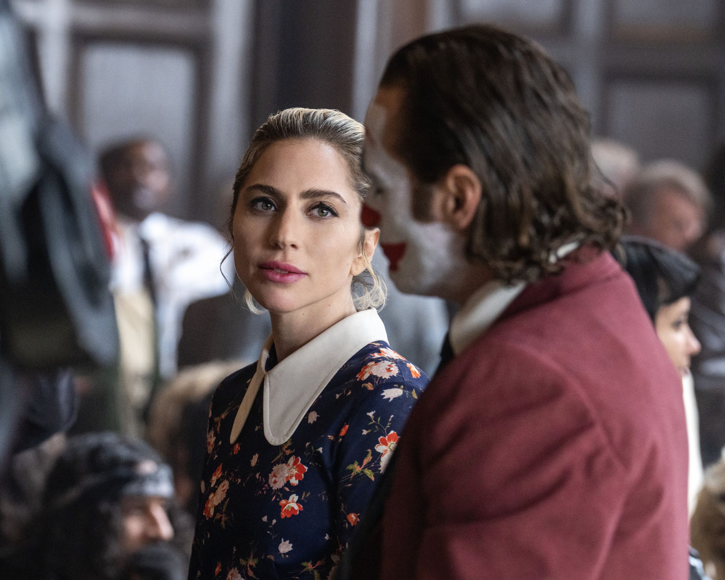 A woman in a black dress with a white collar standing in a courtroom looking at a man in a red suit and clown makeup.