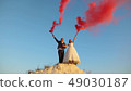 Happy bride and groom waving colored pink smoke against blue sky and laughing. Honeymoon. Romance 49030187