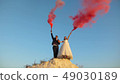 Happy bride and groom waving colored pink smoke against blue sky and laughing. Honeymoon. Romance 49030189