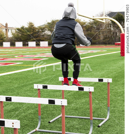 High school athlete jumping over hurdles on a turf field 76479903