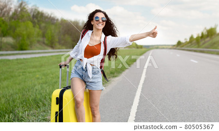 Young lady thumbing her ride, standing with suitcase on highway, trying to catch passing car, hitchhiking in countryside 80505367