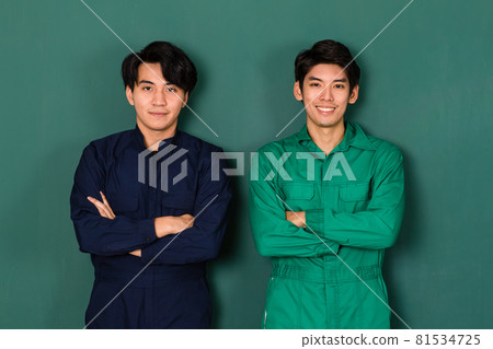 portrait of two smart and handsome young Asian men dressed in blue and green mechanic coveralls stand confidently with crossed arms over green background 81534725