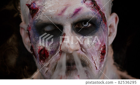 Zombie man with makeup with wounds scars and white contact lenses blows smoke from mouth, smiling 82536295