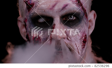 Zombie man with makeup with wounds scars and white contact lenses blows smoke from mouth, smiling 82536296