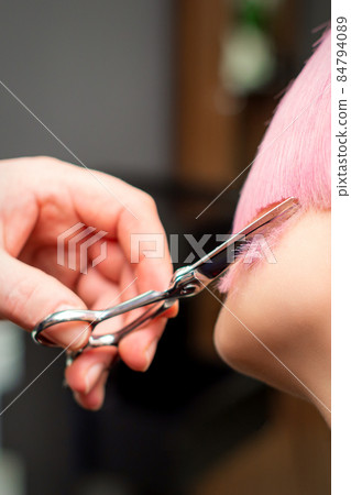 Cutting female hair. Hand of a hairdresser cutting short pink hair of young white woman at the hair salon. 84794089