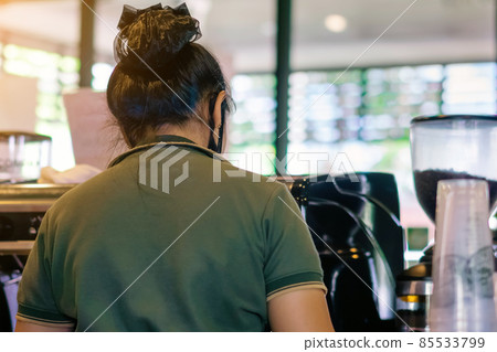 Back view of barista girl prepares coffee in cafe wearing surgical mask due to the pandemic of coronavirus (Covid-19). Young female barista in face mask and standing behind bar counter in coffee shop. 85533799