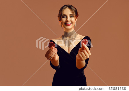 Brunette woman in black dress and sparkling jewelry. She smiling, showing two red chips, posing on brown background. Poker, casino. Close-up 85881388