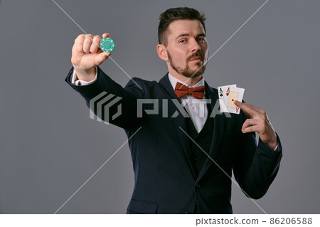 Man in black classic suit and red bow-tie showing two playing cards and chip, posing on gray studio background. Gambling, poker, casino. Close-up. 86206588