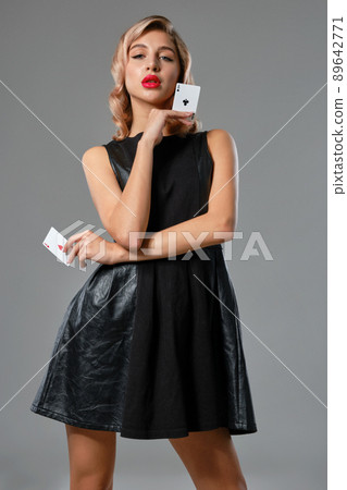 Blonde girl in black stylish dress showing two playing cards, posing against gray background. Gambling entertainment, poker, casino. Close-up. 89642771