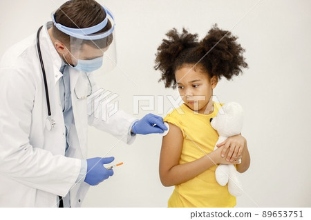 Male doctor in white medical gown is going to do vaccination. Man wearing white medical gown and face mask. Girl wearing yellow t-shirt and holding a bear toy in hands. 89653751