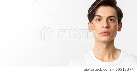 People, lgbtq and beauty concept. Close-up of beautiful androgynous man with glitter on face, looking at camera, standing over white background 90512374