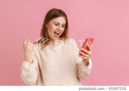 Extremely happy excited blond woman standing with cell phone in hands and clenching fist, celebrating her victory, great news, wearing white sweater. Indoor studio shot isolated on pink background. 91047228