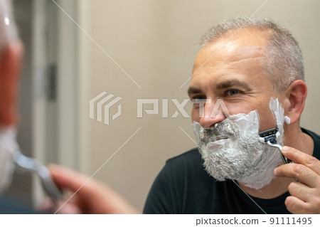 Mature gray-haired man restyling his beard himself at home using razor 91111495