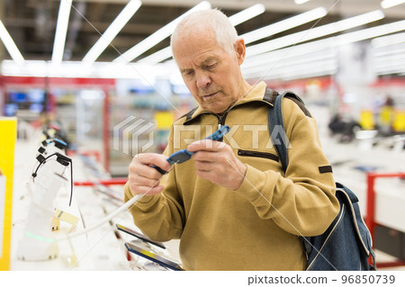 elderly grayhaired man pensioner examining counter with electronic gadgets and smart watches in showroom of digital goods store 96850739