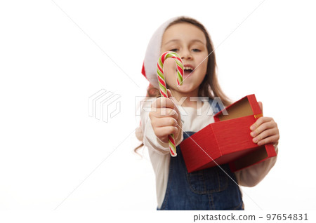 Focus on red green striped lillipop in the hands of a blurred overjoyed happy smiling child girl in Santa hat, with gift box, holding out a sweet candy cane to camera, isolated over white background 97654831