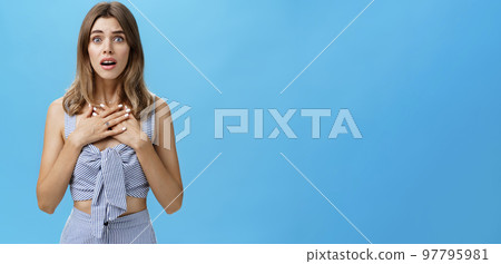 Waist-up shot of woman feeling bad for friend empathizing hearing shocking story gasping opening mouth and looking speechless holding palms on breast standing intense and nervous over blue wall 97795981