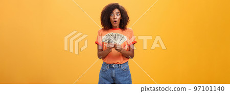 Woman shocked finding lots of cash in safe. Portrait of surprised speechless good-looking dark-skinned female with curly haircut folding lips gasping holding money posing over orange background 97101140