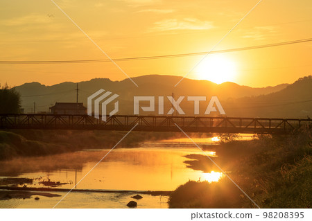 The morning sun rising over the mountain village and the river reflecting the sunrise 98208395