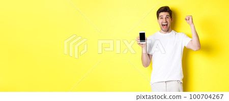 Cheerful guy showing smartphone screen, raising hand up and celebrating, triumphing over internet achievement, standing over yellow background 100704267