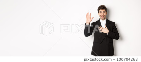 Image of handsome smiling guy telling the truth, raising on arm to promise something, standing over white background in black suit 100704268