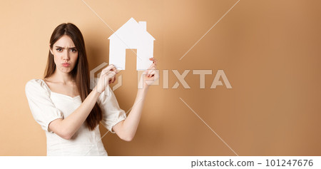 Real estate. Angry girl showing house cutout and frowning upset, complaining on apartment, standing against beige background 101247676