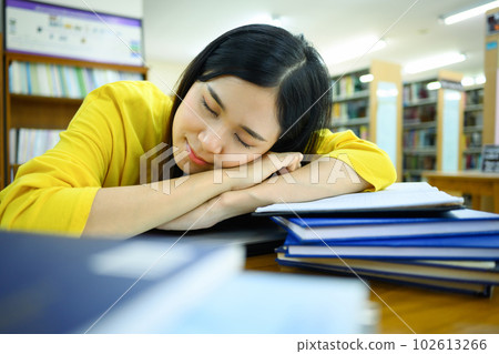 Tired female student sleeping on pile of books in the library with blurred bookshelf on background 102613266