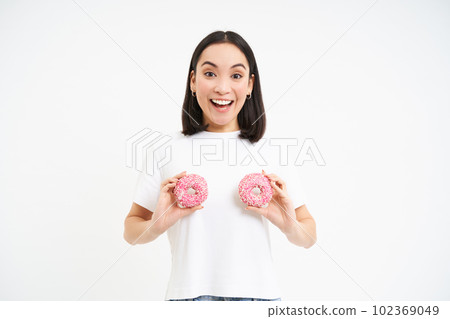 Smiling happy asian woman laughing, holding two glazed doughnuts over chest, standing over white background 102369049