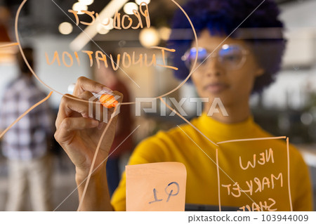 Biracial casual businesswoman with blue afro making notes on glass wall in office 103944009