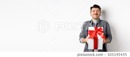 Happy handsome young man holding big valentines day gift and cheering, receive romantic surprise present from lover, standing on white background 103145435