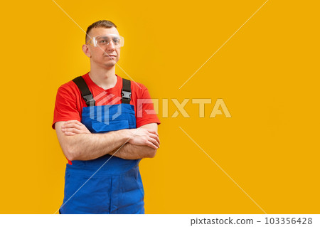 Serious middle aged engineer wearing blue overalls crossed arms standing isolated on yellow background. Copy space, mockup 103356428