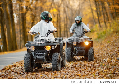 A young caucasian man and woman riding an ATV quad bikes in autumn forest. Two friends maneuvering off-road ATV. Couple wearing grey sportive costumes. 105469883