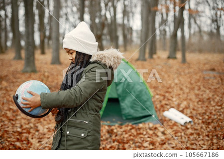 One little black girl in tent camping in the forest. Girl is holding a globe in her hands. Black girl wearing khaki coat and beige hat. 105667186