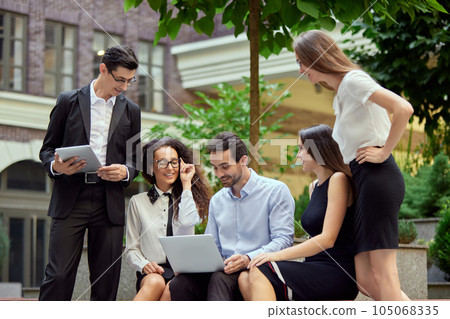 Group of employees, business people, project leaders sitting on bench outside office on daytime and working online on laptop 105068335