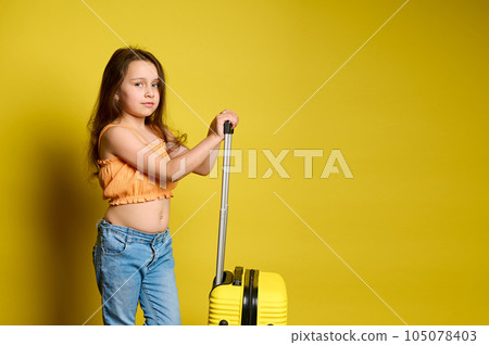 Beautiful little traveler girl with long hair, dressed in blue jeans and orange top, with suitcase, yellow background. 105078403