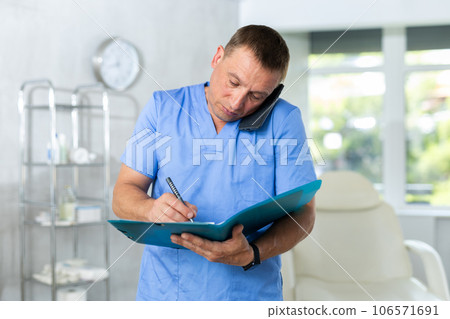 Middle-aged man doctor standing in doctor's cabinet with documents in hands 106571691