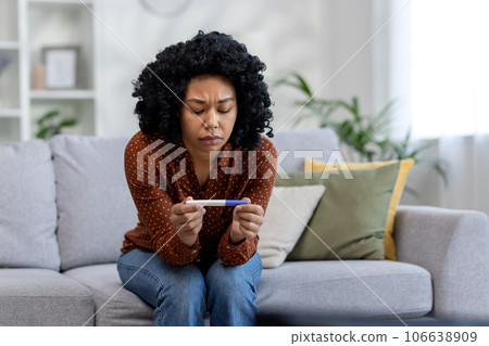 Upset and pensive African American young woman sitting on sofa at home and looking at negative pregnancy test. Ovulation test. 106638909