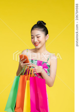 Young energetic Asian woman holding mobile phone with shopping bags on yellow background 106784988