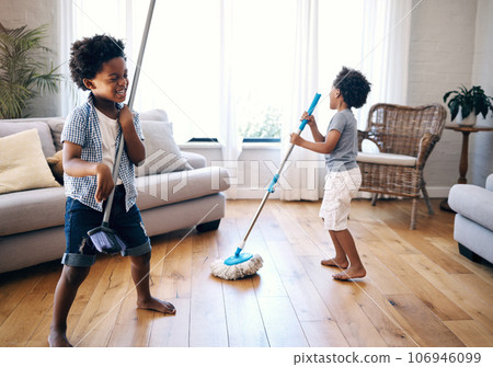 Two mixed race little boys playing with a mop and broom in the lounge at home. Happy siblings having fun playing with cleaning supplies at home. Carefree brothers singing while cleaning at home 106946099