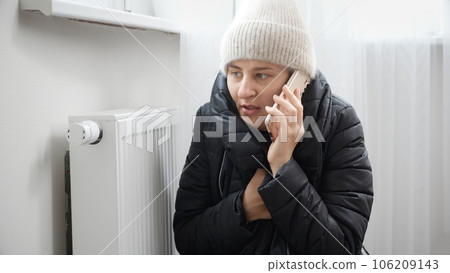 Portrait of stressed brunette woman in winter coat and woolen hat calling service to repair broken heater radiator 106209143