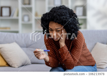 Upset young African American woman sitting on sofa at home, holding hand to head and looking shocked at quick pregnancy test with negative result. 106213978