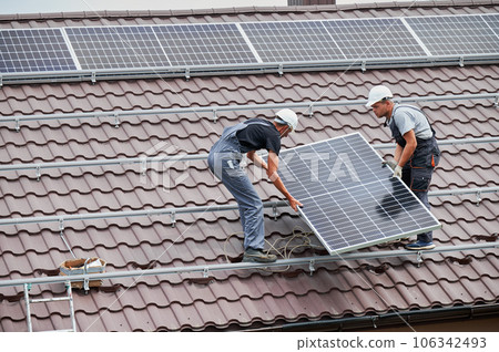 Men workers carrying photovoltaic solar moduls on roof of house. Electricians in helmets installing solar panel system outdoors. Concept of alternative and renewable energy. 106342493