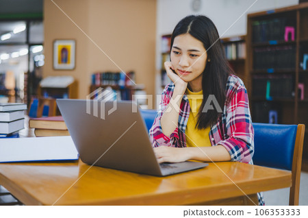 Female college student working on laptop and searching for books to study, make report, find useful information in college room. Concept of reading, learning and intelligence. 106353333